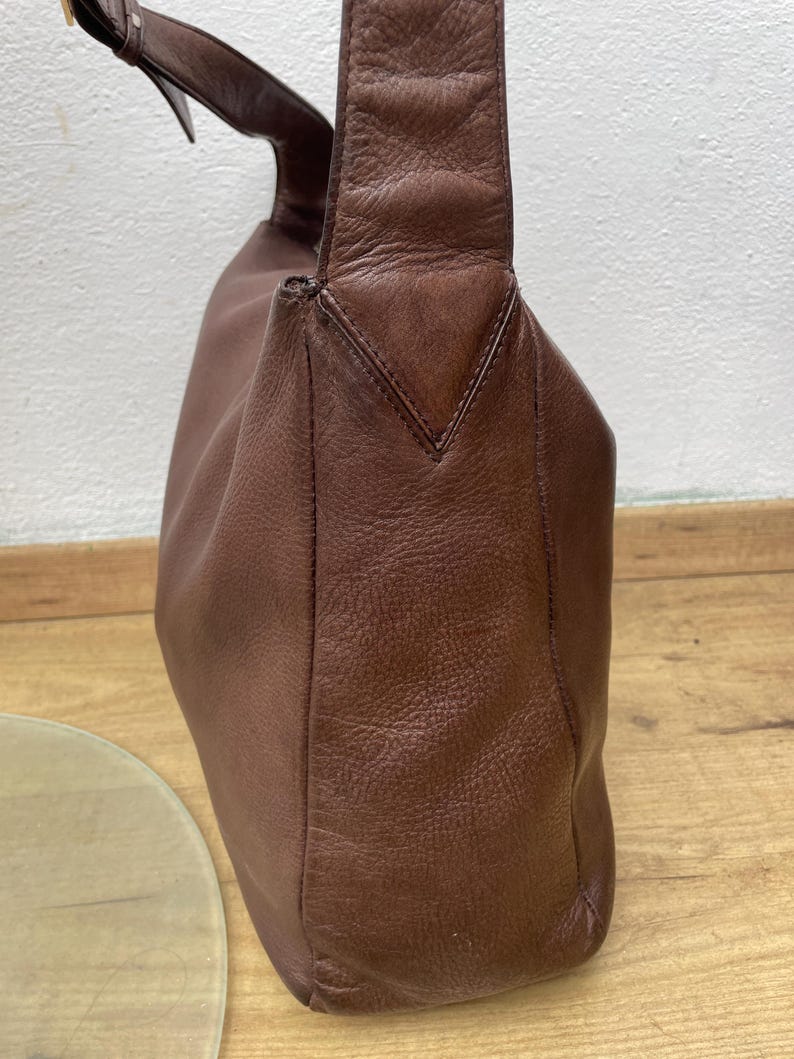 May include: A brown leather shoulder bag with a single strap and a buckle closure. The bag has a rounded shape and a textured surface. The strap is attached to the bag with a metal buckle. The bag is sitting on a wooden surface.