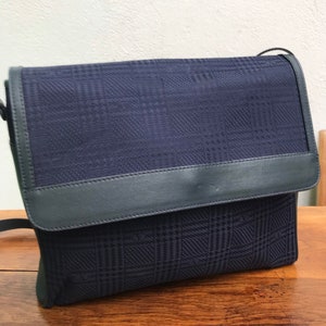 May include: A navy blue crossbody bag with a plaid pattern. The bag features a flap closure and a dark blue leather trim. The bag is sitting on a wooden surface.