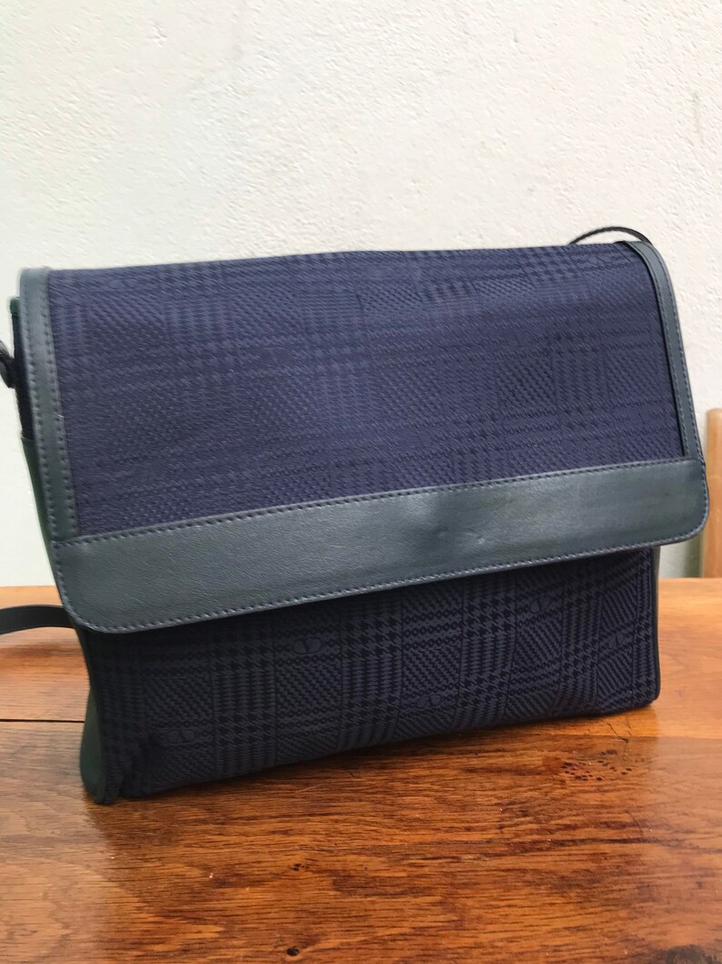 May include: A navy blue crossbody bag with a plaid pattern. The bag features a flap closure and a dark blue leather trim. The bag is sitting on a wooden surface.
