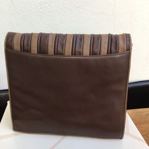 May include: A rectangular clutch in shades of brown. The main body is a deep chocolate brown, with a lighter tan accent on the top. The top section features vertical pleats, adding texture to the design. The clutch is resting on a white surface.
