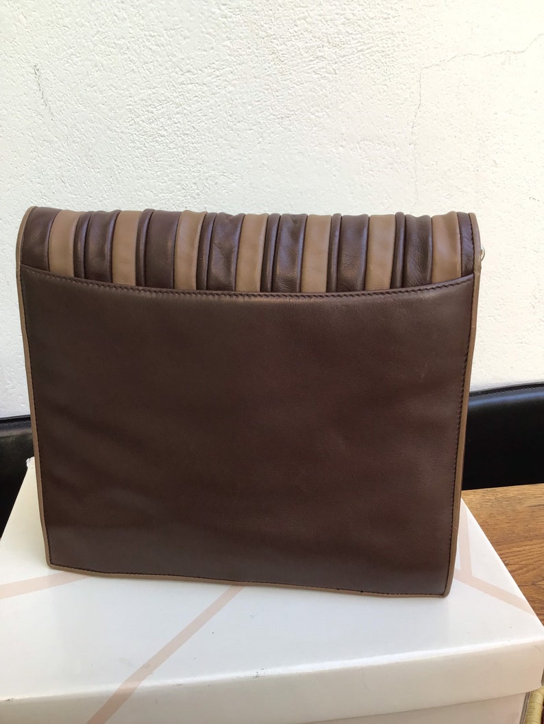 May include: A rectangular clutch in shades of brown. The main body is a deep chocolate brown, with a lighter tan accent on the top. The top section features vertical pleats, adding texture to the design. The clutch is resting on a white surface.