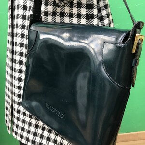 Puede incluir: Un bolso de hombro de charol azul marino oscuro con acabado brillante, que presenta un cierre de solapa, una hebilla de metal dorado en la correa lateral y la palabra VALENTINO grabada en letras may&uacute;sculas en la parte inferior frontal.