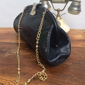 May include: A vintage black handbag with a gold chain strap. The bag features a textured, reptile-like pattern and a clasp closure. The gold chain is attached with small gold accents. The bag is on a wooden surface. The bag is a classic design.