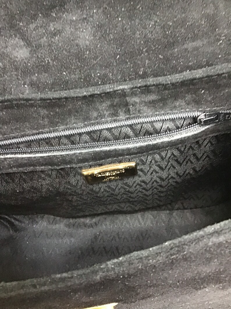 May include: Black suede handbag with a zippered closure. The interior features a patterned lining and a gold-toned label. The bag's design is simple and elegant, suitable for various occasions.
