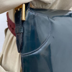 Puede incluir: Un bolso de hombro de charol en color verde azulado oscuro con acabado brillante y una hebilla de metal dorado en la correa lateral. El bolso tiene un dise&ntilde;o estructurado con costuras visibles en los bordes donde la correa se une al cuerpo.