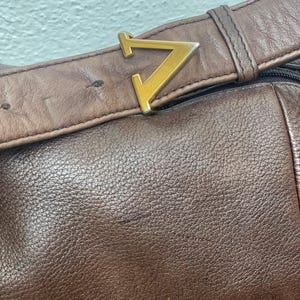 May include: Close-up of a brown leather bag with a gold-colored metal buckle in the shape of a stylized letter "V". The leather has a textured surface and the bag appears to be well-worn.