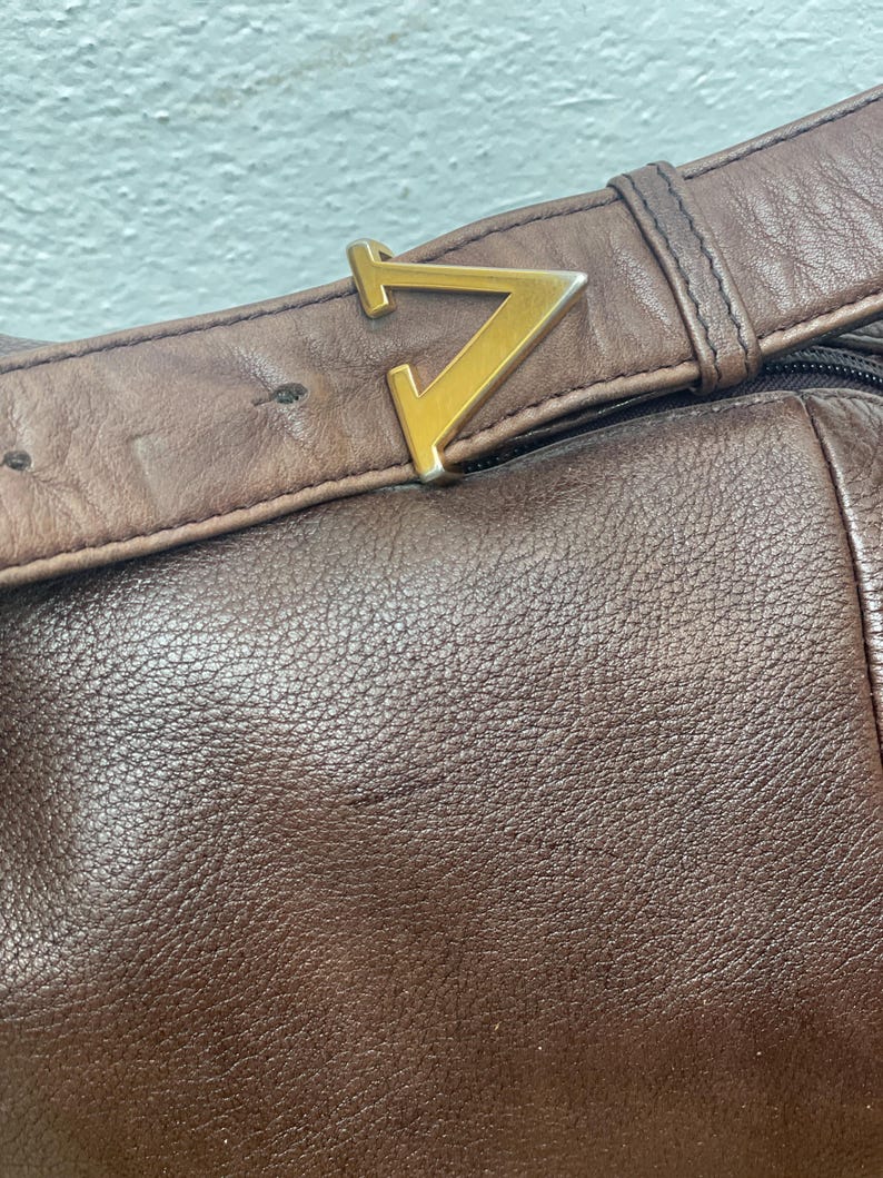 May include: Close-up of a brown leather bag with a gold-colored metal buckle in the shape of a stylized letter "V". The leather has a textured surface and the bag appears to be well-worn.