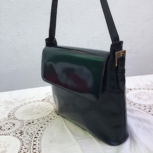 Puede incluir: Un bolso de hombro de charol negro con acabado brillante, que presenta un cierre de solapa rectangular, una correa de hombro &uacute;nica ajustable y un detalle de hebilla de metal dorado en el punto de fijaci&oacute;n lateral de la correa.