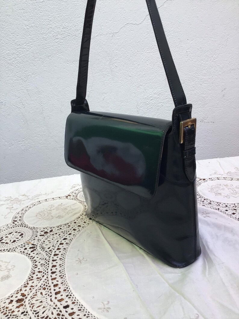 Puede incluir: Un bolso de hombro de charol negro con acabado brillante, que presenta un cierre de solapa rectangular, una correa de hombro &uacute;nica ajustable y un detalle de hebilla de metal dorado en el punto de fijaci&oacute;n lateral de la correa.