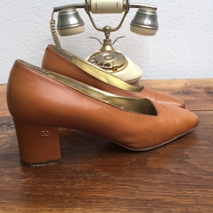 May include: A pair of brown leather pumps with a low block heel. The shoes have a pointed toe and a gold trim along the inner edge. The heels have a CD logo. An antique telephone is in the background.