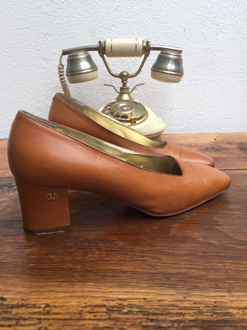 May include: A pair of brown leather pumps with a low block heel. The shoes have a pointed toe and a gold trim along the inner edge. The heels have a CD logo. An antique telephone is in the background.