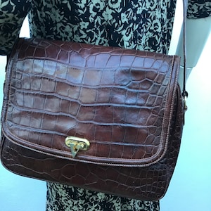 Op de afbeelding: Een bruine leren crossbodytas met een krokodillenleertextuur. De tas heeft een rechthoekige vorm met een klepsluiting en een goudkleurige sluiting. De riem is dun en bruin. De tas wordt gedragen over een jurk met bloemenpatroon.
