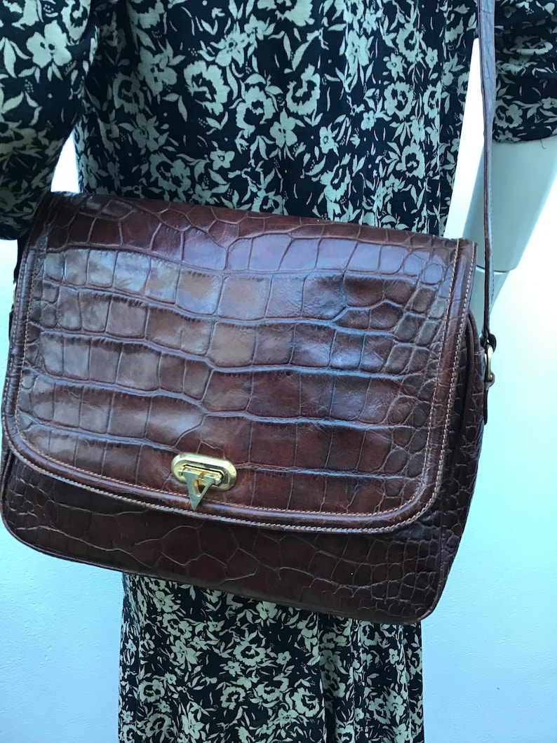 Op de afbeelding: Een bruine leren crossbodytas met een krokodillenleertextuur. De tas heeft een rechthoekige vorm met een klepsluiting en een goudkleurige sluiting. De riem is dun en bruin. De tas wordt gedragen over een jurk met bloemenpatroon.
