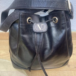 May include: Black leather bucket bag with a drawstring closure and a shoulder strap. The bag features a gold-tone buckle and a silver-tone logo on the front. The bag has a textured surface and a rounded shape.