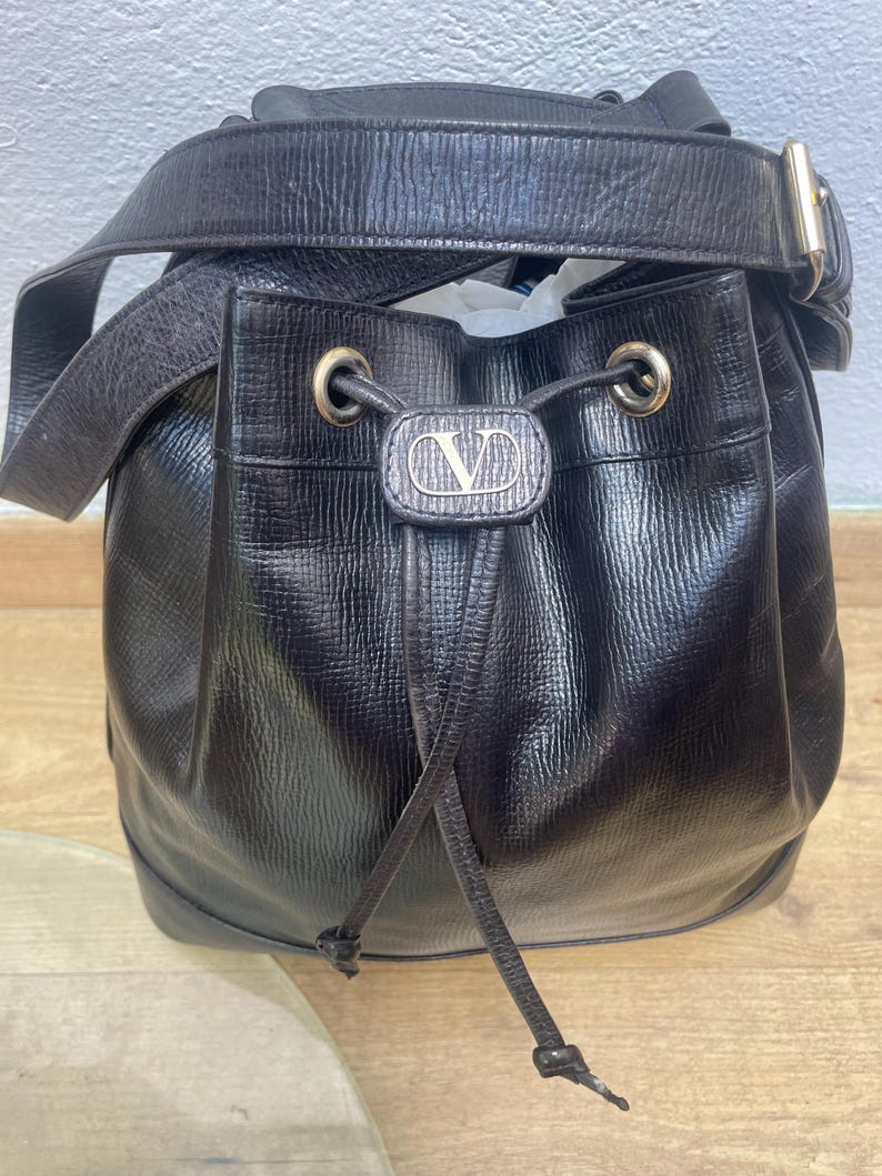 May include: Black leather bucket bag with a drawstring closure and a shoulder strap. The bag features a gold-tone buckle and a silver-tone logo on the front. The bag has a textured surface and a rounded shape.