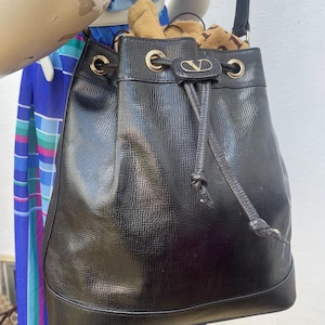 May include: Black leather bucket bag with a drawstring closure and gold-tone hardware. The bag features a textured surface and a small logo detail. The strap is black and the bag is filled with items.