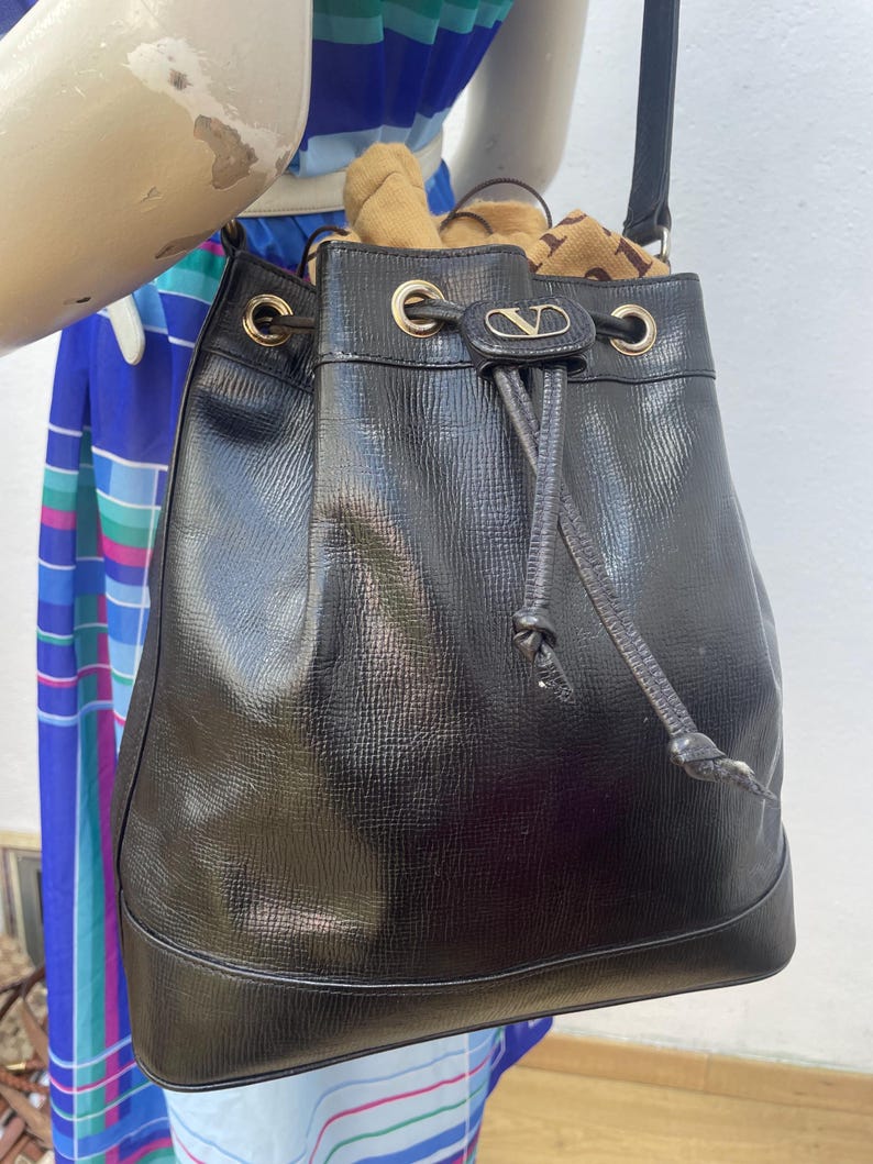 May include: Black leather bucket bag with a drawstring closure and gold-tone hardware. The bag features a textured surface and a small logo detail. The strap is black and the bag is filled with items.