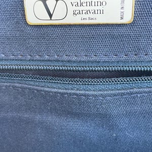 May include: Close-up of a navy blue fabric with a zipper and a rectangular label. The label has a gold border and features the Valentino Garavani logo and text. The text "Les Sacs" is also visible.