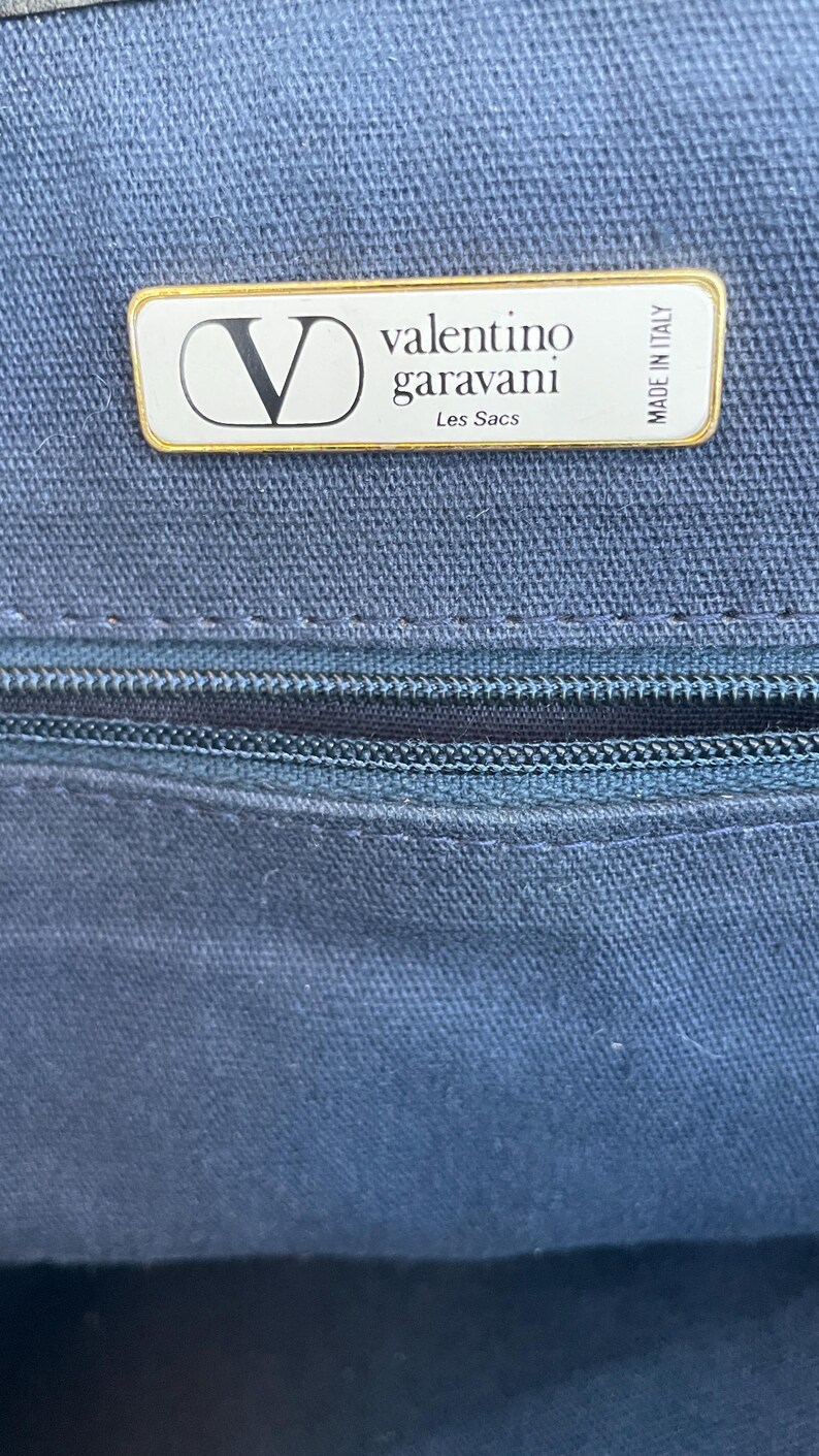 May include: Close-up of a navy blue fabric with a zipper and a rectangular label. The label has a gold border and features the Valentino Garavani logo and text. The text "Les Sacs" is also visible.
