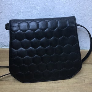 May include: Black leather crossbody bag with a geometric honeycomb pattern on the front. The bag features a long, thin strap and a rectangular shape with rounded corners. The bag is on a wooden surface.
