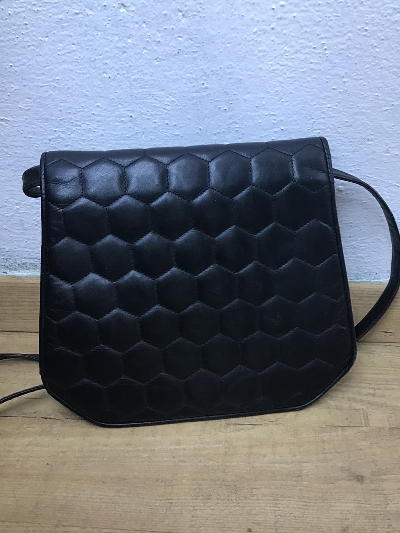 May include: Black leather crossbody bag with a geometric honeycomb pattern on the front. The bag features a long, thin strap and a rectangular shape with rounded corners. The bag is on a wooden surface.