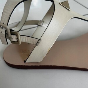 May include: Off-white leather sandals with a flat sole. The sandals feature a buckle closure and are adorned with metallic pyramid studs. The design includes a thong strap and ankle strap for secure wear.