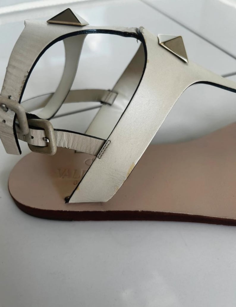 May include: Off-white leather sandals with a flat sole. The sandals feature a buckle closure and are adorned with metallic pyramid studs. The design includes a thong strap and ankle strap for secure wear.