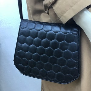 May include: Black shoulder bag with a geometric honeycomb pattern. The bag has a flap closure and a long, thin strap. The bag is made of a smooth material. The bag is hanging on a mannequin.