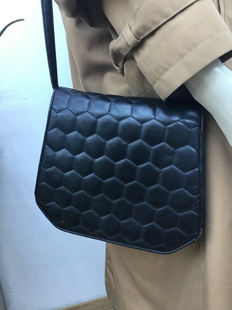 May include: Black shoulder bag with a geometric honeycomb pattern. The bag has a flap closure and a long, thin strap. The bag is made of a smooth material. The bag is hanging on a mannequin.