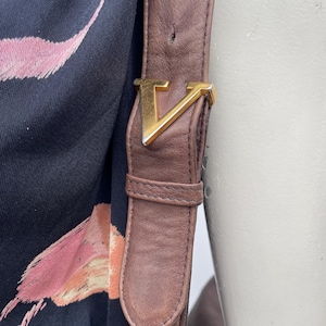 May include: Close-up of a brown leather shoulder bag strap with a gold-colored metal buckle in the shape of a "V". The strap is attached to a black fabric with a pink and orange floral pattern.