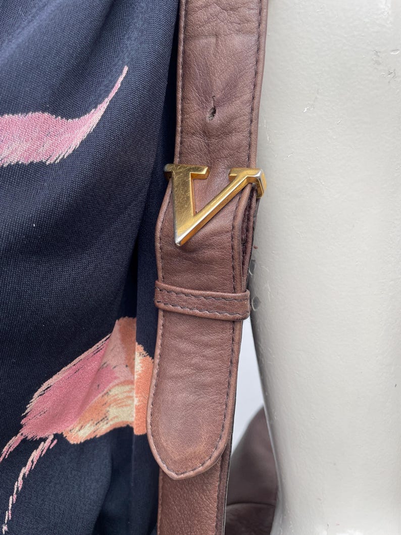 May include: Close-up of a brown leather shoulder bag strap with a gold-colored metal buckle in the shape of a "V". The strap is attached to a black fabric with a pink and orange floral pattern.