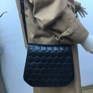 May include: Black leather crossbody bag featuring a geometric honeycomb design. The bag has a long, adjustable strap and a flap closure. The bag is worn over a tan trench coat.
