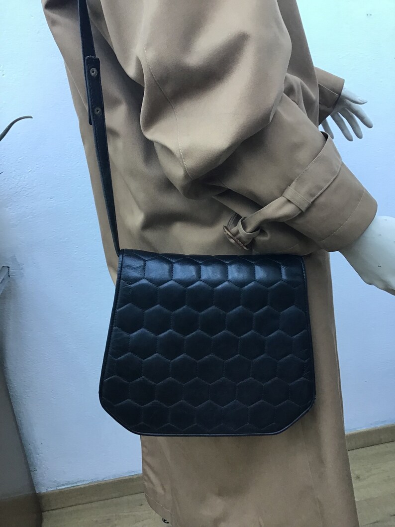 May include: Black leather crossbody bag featuring a geometric honeycomb design. The bag has a long, adjustable strap and a flap closure. The bag is worn over a tan trench coat.
