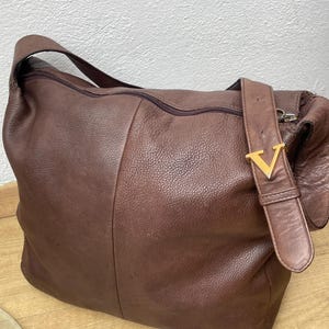 May include: A brown leather shoulder bag with a zipper closure and a strap. The bag has a gold-colored metal buckle with a "V" design. The leather has a textured appearance.