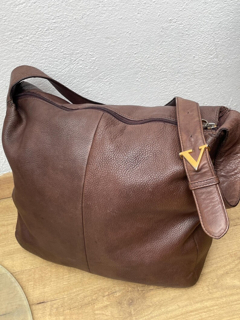 May include: A brown leather shoulder bag with a zipper closure and a strap. The bag has a gold-colored metal buckle with a "V" design. The leather has a textured appearance.