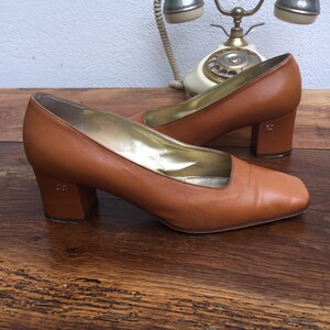 May include: A pair of brown leather pumps with a square toe and block heel. The interior is gold-colored. The heels have a small logo. The shoes are on a wooden surface with a vintage telephone in the background.