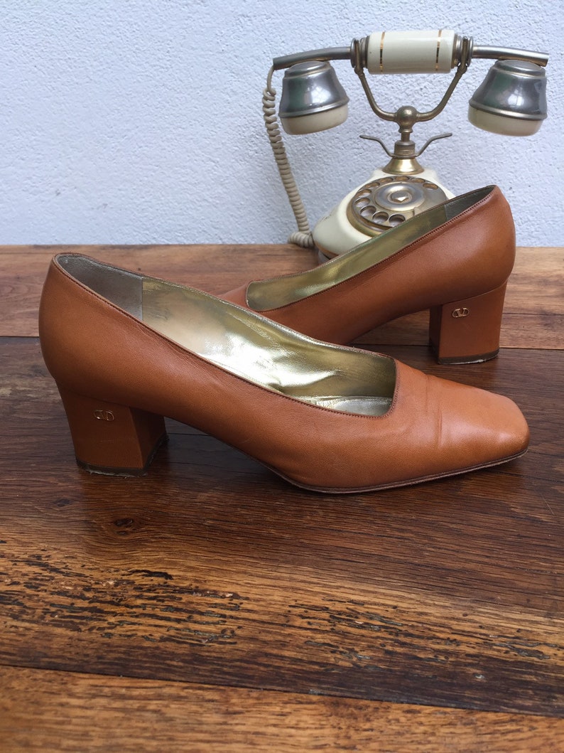 May include: A pair of brown leather pumps with a square toe and block heel. The interior is gold-colored. The heels have a small logo. The shoes are on a wooden surface with a vintage telephone in the background.