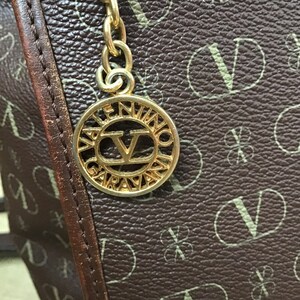 K&ouml;nnte beinhalten: Eine braune Ledertasche mit einer goldfarbenen Kette und einem runden Anh&auml;nger mit der Aufschrift "Valentino Garavani" und einem gro&szlig;en "V" in der Mitte.