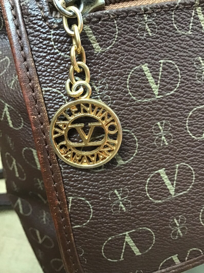 K&ouml;nnte beinhalten: Eine braune Ledertasche mit einer goldfarbenen Kette und einem runden Anh&auml;nger mit der Aufschrift "Valentino Garavani" und einem gro&szlig;en "V" in der Mitte.