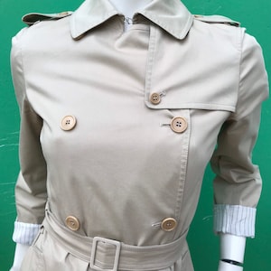 May include: A beige trench coat with a double-breasted design, featuring large, round buttons and a matching belt with a square buckle. The coat has a classic collar and epaulettes, with the sleeves rolled up to reveal a striped lining.