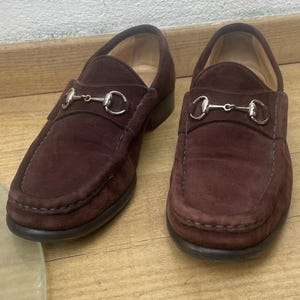 Peut inclure: Une paire de mocassins en daim marron foncé avec un détail de mors en métal argenté sur le dessus. Les chaussures présentent un bout arrondi, des coutures apparentes le long de l'empeigne et un petit talon, dans un style à enfiler classique.