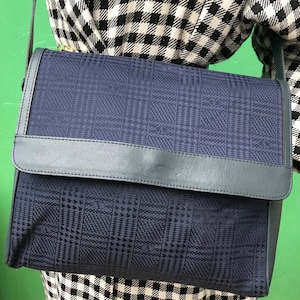 May include: A navy blue shoulder bag with a flap closure. The bag features a plaid pattern and a leather trim. The bag is being worn over a black and white checkered outfit.
