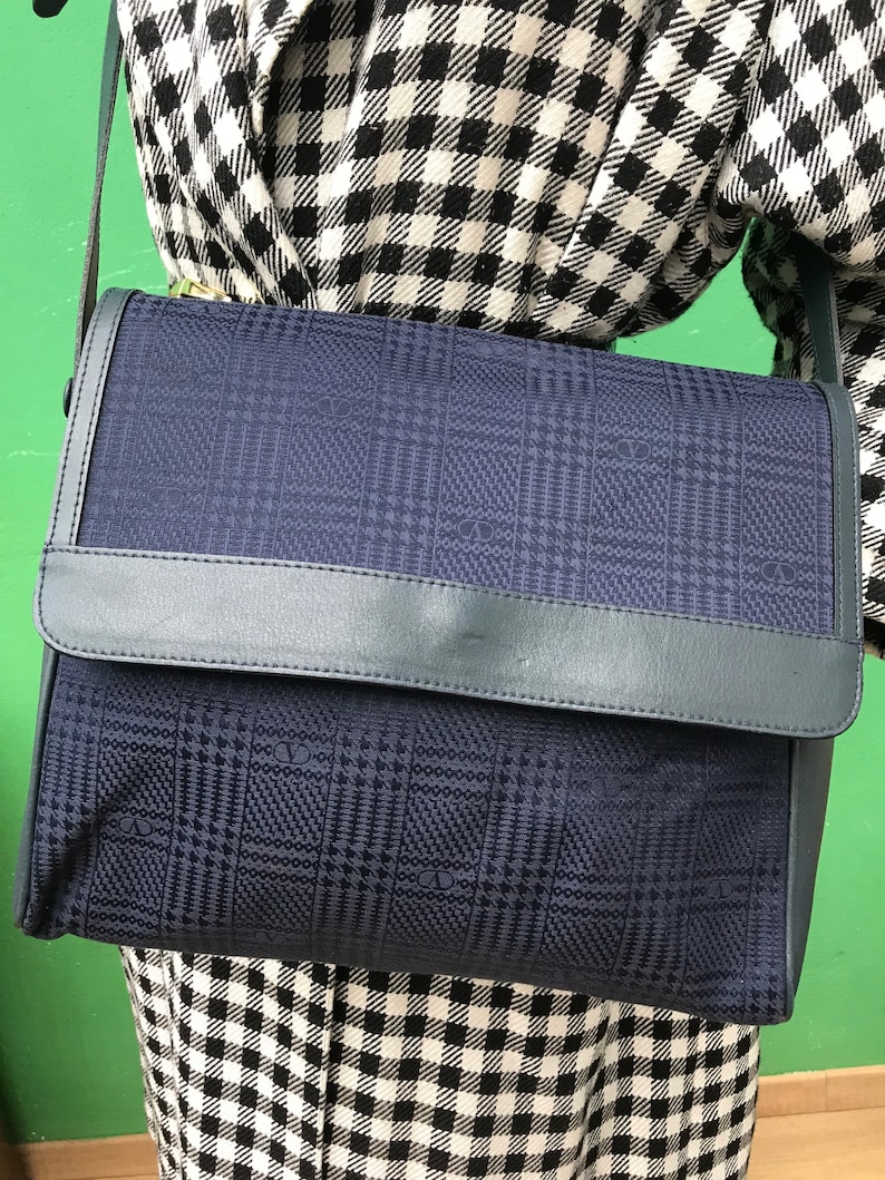May include: A navy blue shoulder bag with a flap closure. The bag features a plaid pattern and a leather trim. The bag is being worn over a black and white checkered outfit.