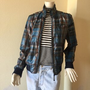 May include: A plaid bomber jacket in shades of blue and brown, with a black zipper, is displayed on a mannequin. The jacket is paired with a black and white striped shirt and light blue jeans. The jacket has a ribbed collar and cuffs.