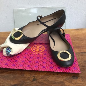 May include: Black leather flats with a gold-tone circular buckle. The shoes have a strap across the top of the foot and are displayed on a pink and orange patterned box. A white cloth bag is also present.