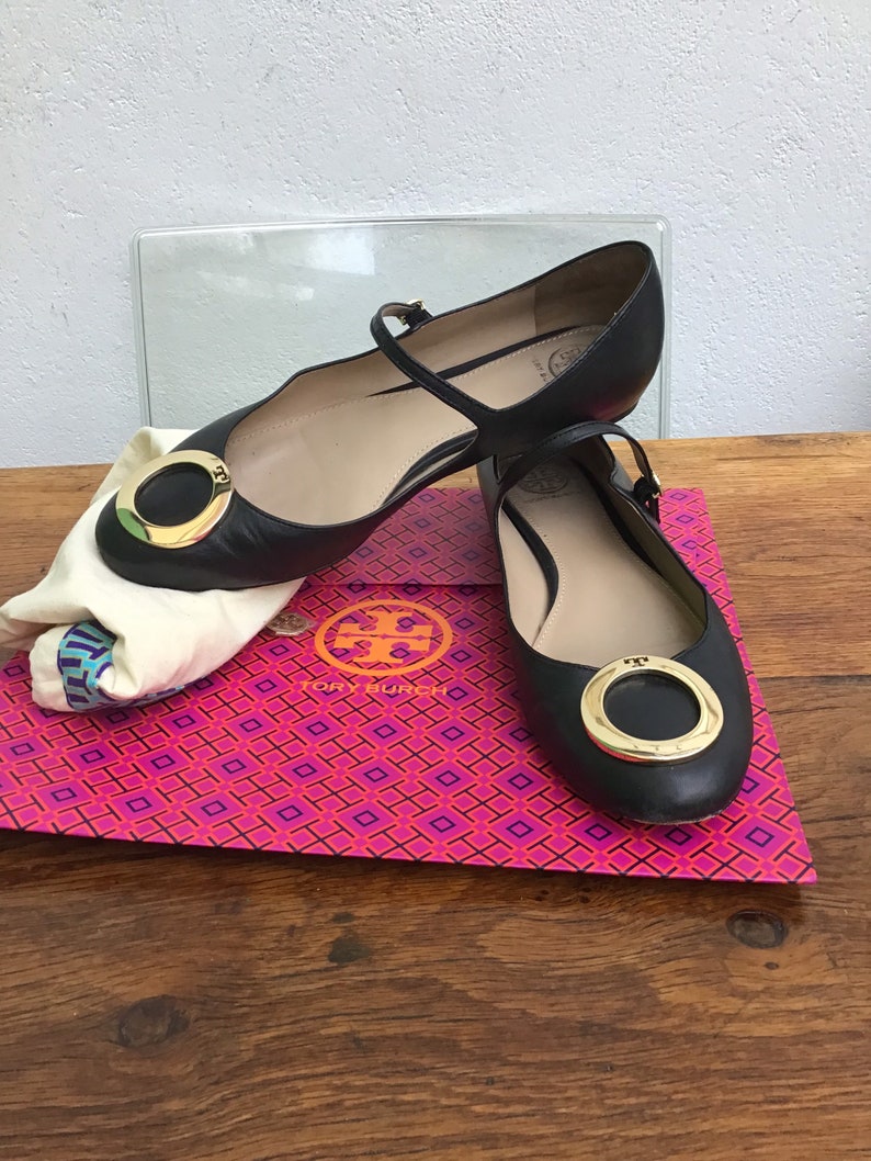 May include: Black leather flats with a gold-tone circular buckle. The shoes have a strap across the top of the foot and are displayed on a pink and orange patterned box. A white cloth bag is also present.