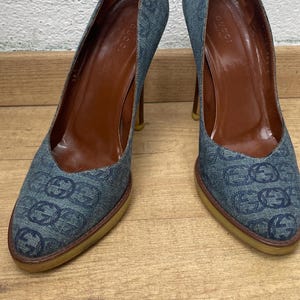Peut inclure: Une paire d'escarpins en jean avec un motif de logo répété. Les talons ont un bout pointu, une semelle intérieure en cuir marron et un talon aiguille. Les chaussures sont présentées sur une surface en bois.