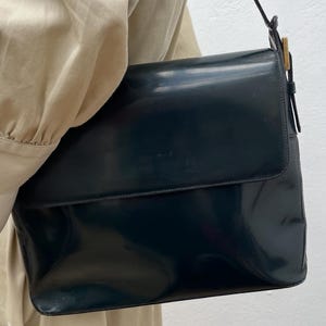 Puede incluir: Un bolso de hombro de color azul marino oscuro con un acabado liso y brillante y un cierre de solapa rectangular. El bolso cuenta con una correa fina a juego sujeta con herrajes met&aacute;licos de tono dorado en el lateral, lo que le confiere un aspecto estructurado.