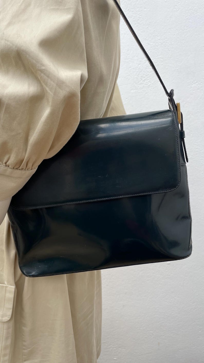 Puede incluir: Un bolso de hombro de color azul marino oscuro con un acabado liso y brillante y un cierre de solapa rectangular. El bolso cuenta con una correa fina a juego sujeta con herrajes met&aacute;licos de tono dorado en el lateral, lo que le confiere un aspecto estructurado.