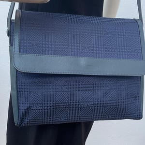 May include: A navy blue shoulder bag with a flap closure. The bag features a plaid pattern and a gray leather trim. The strap is also gray. The bag is displayed on a mannequin wearing a black dress.